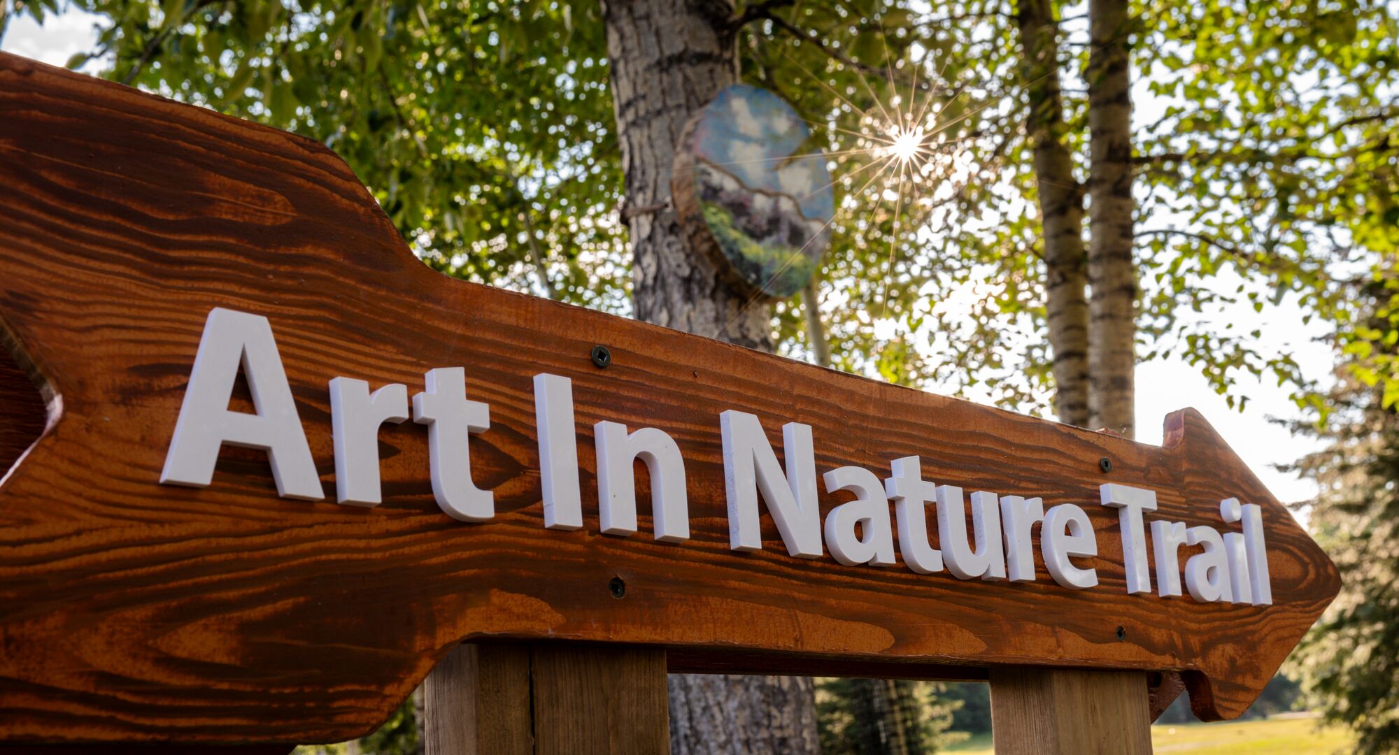 A sign that reads the Art in Nature Trail with an art piece on a tree in the background in Banff National Park.